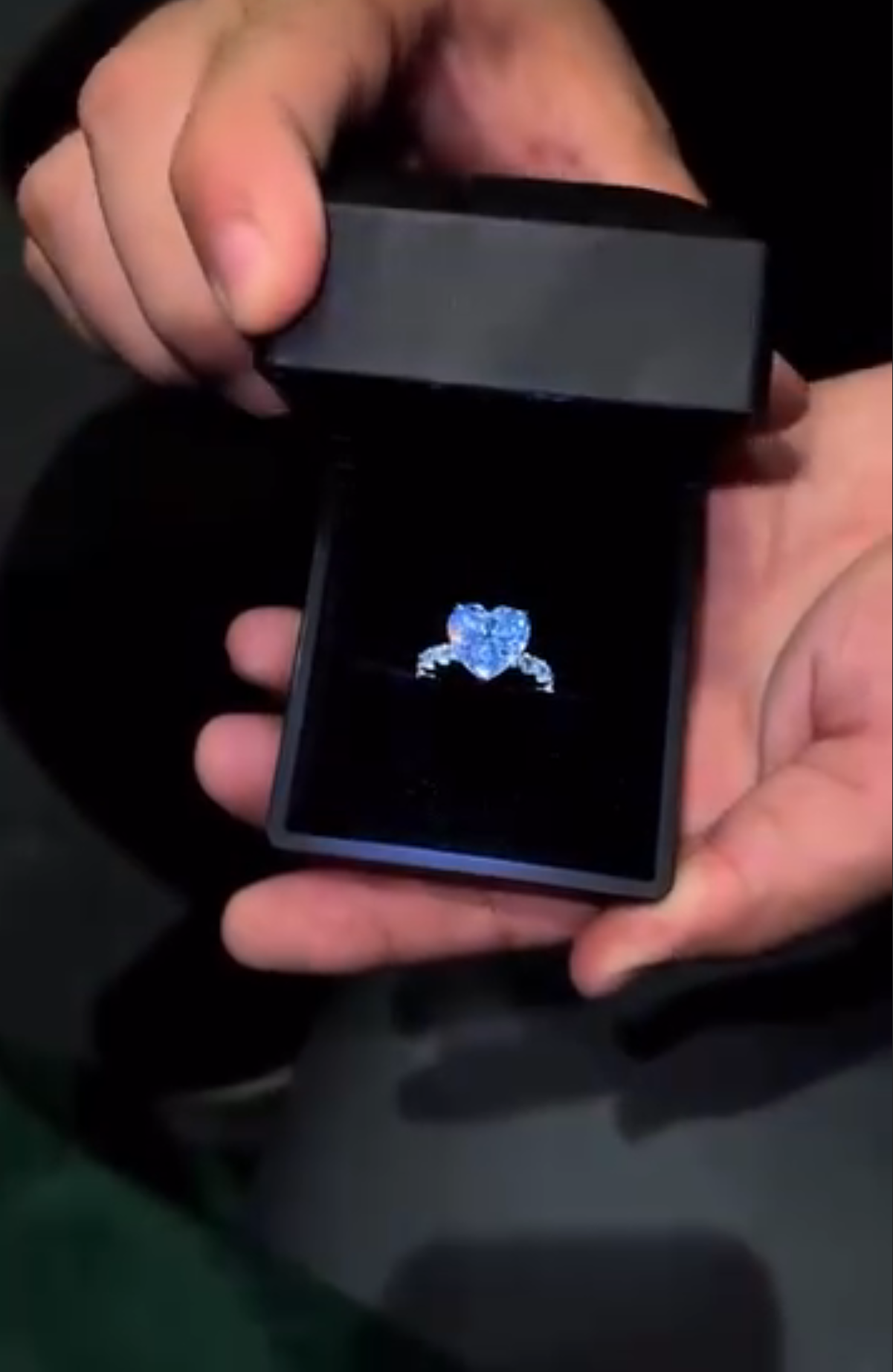 Person holding a black jewelry box with a blue gemstone ring inside on a dark background